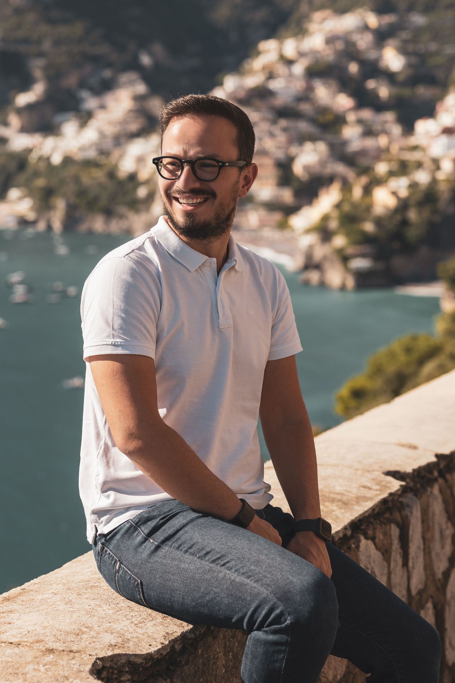 Someone in glasses and a white polo shirt sits on a stone wall overlooking a scenic coastal view with turquoise waters.
