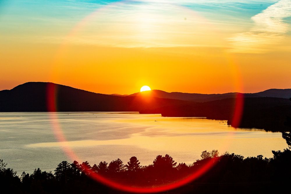 Dramatic golden sunset over a mountain lake with vivid orange sky and lens flare reflection on water.