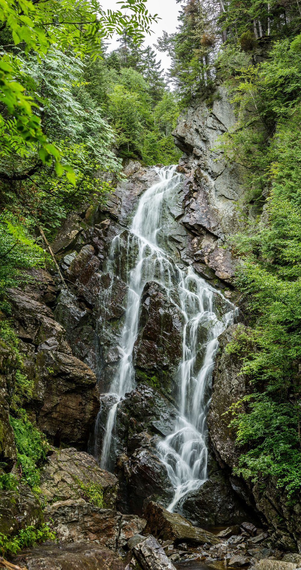Tall cascading waterfall flowing over mossy rocky cliffs surrounded by lush green forest vegetation.