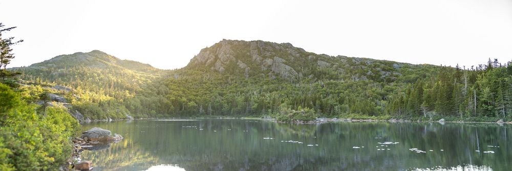 Panoramic view of a tranquil mountain lake with rocky shores and sun-lit forested hills in background.