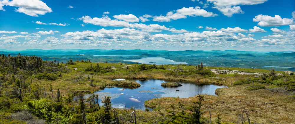 Panoramic highland vista featuring a small reflective pond, rolling green hills, and distant lakes under blue sky.