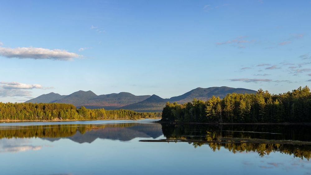 Calm lake at golden hour reflecting forested shores and distant mountain peaks under a clear blue sky.