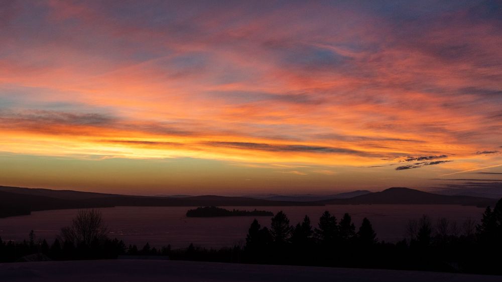 Vibrant orange and red sunset sky over a tranquil lake with silhouetted trees and mountains.