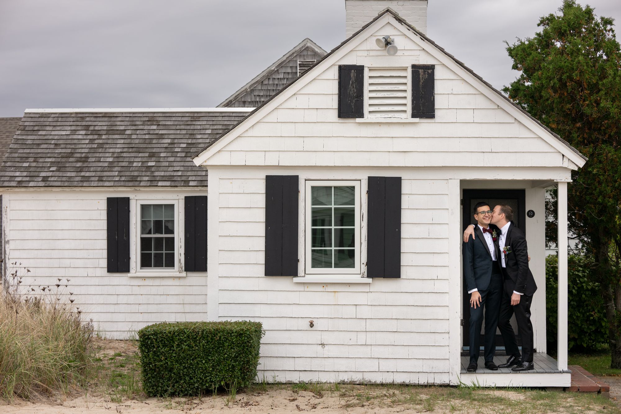 two grooms in tuxedos in the doorway of a white cottage captured by Cape Cod wedding photographer