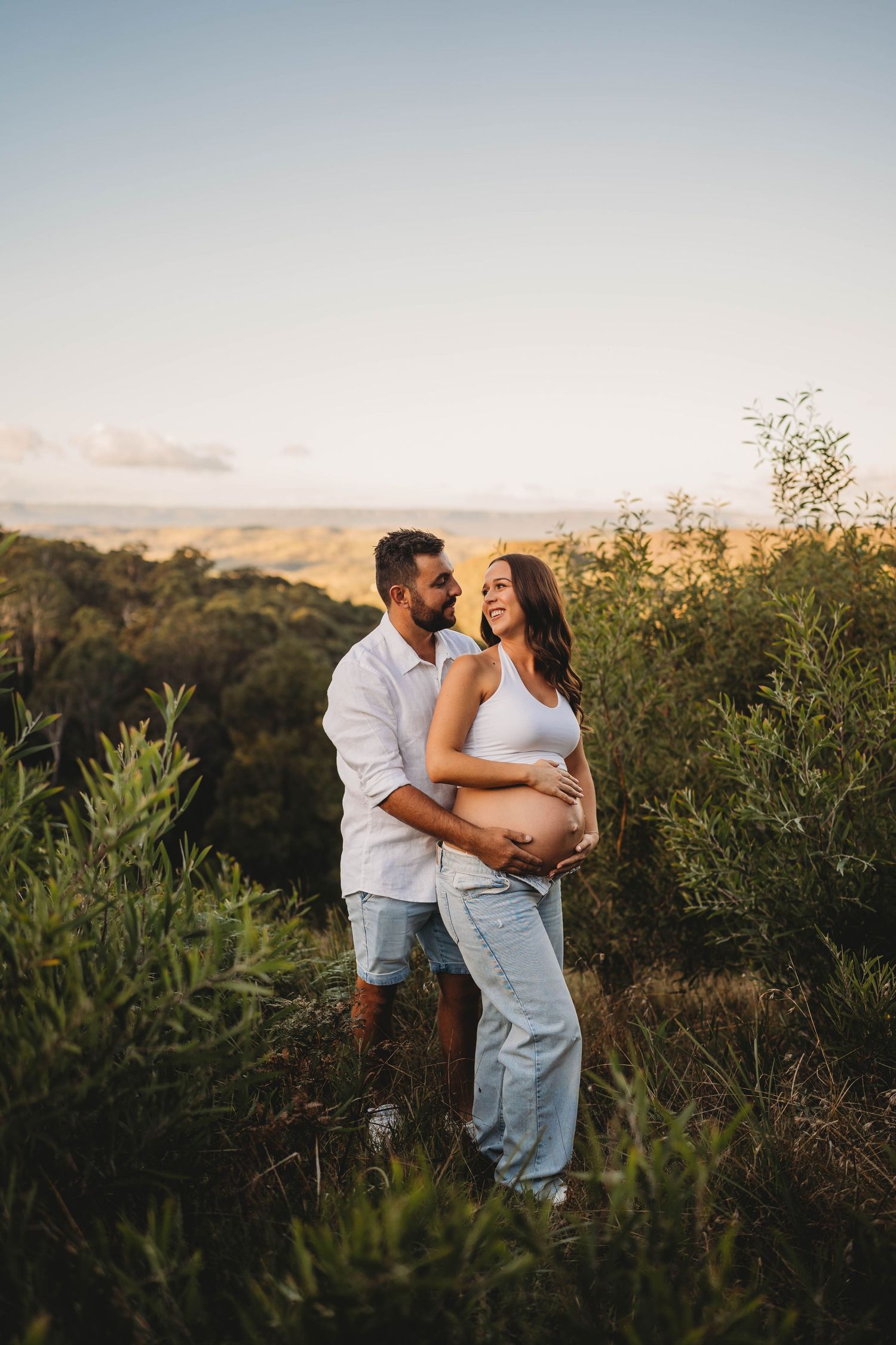 Couple embracing in lush greenery at sunset with woman showing her pregnant belly.