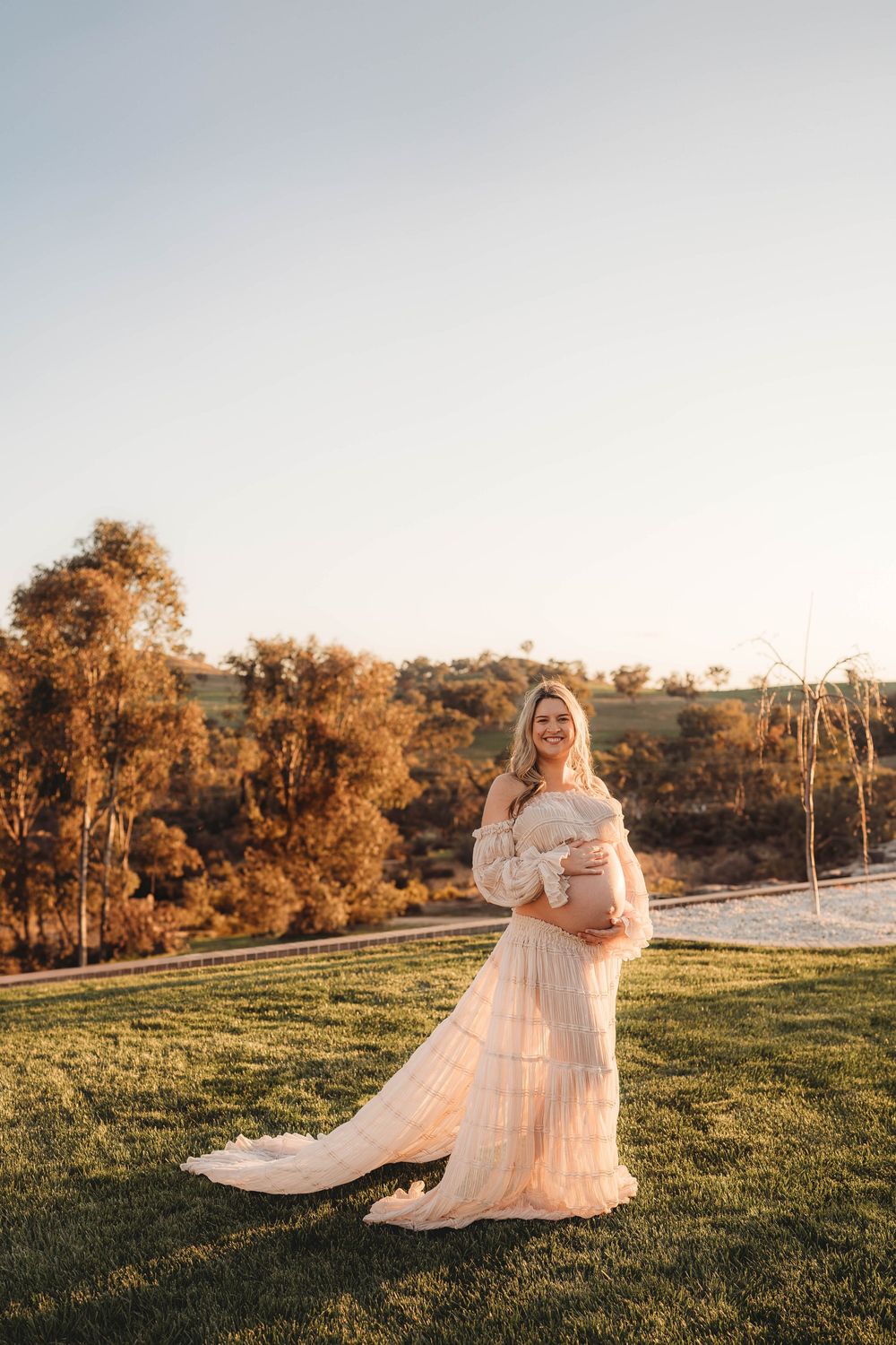 Smiling pregnant woman in elegant off-shoulder cream lace gown standing on green lawn at golden hour.