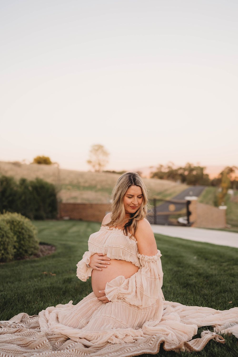 Pregnant woman in off-shoulder boho cream dress sitting on blanket outdoors at golden sunset.