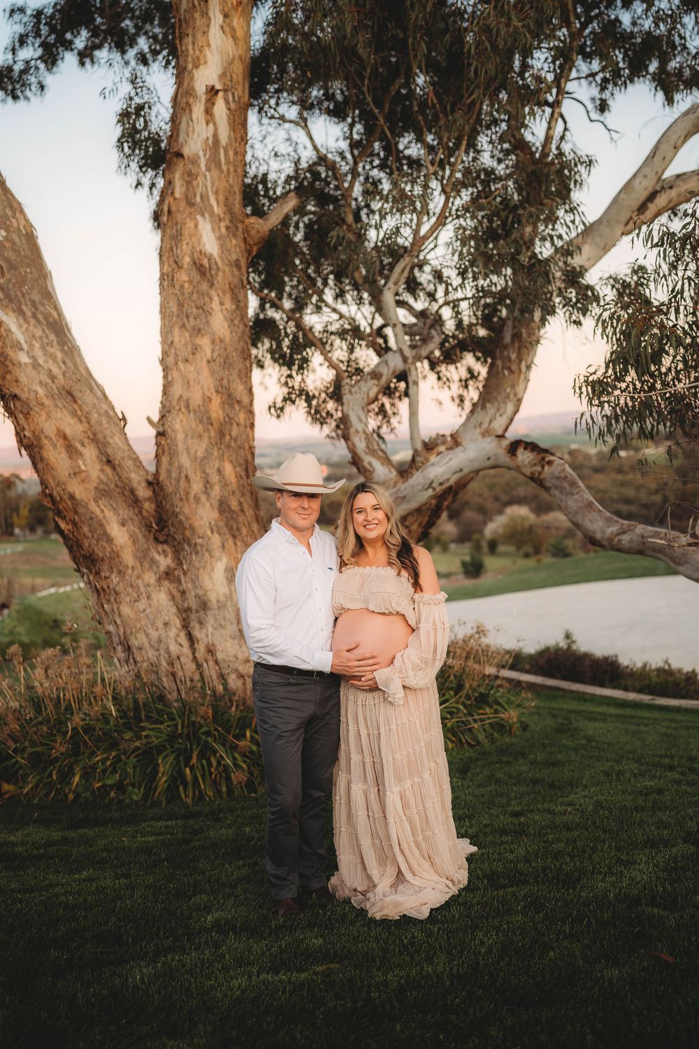 Couple posing under large eucalyptus tree at golden hour, man in cowboy hat embracing pregnant woman in lace dress.
