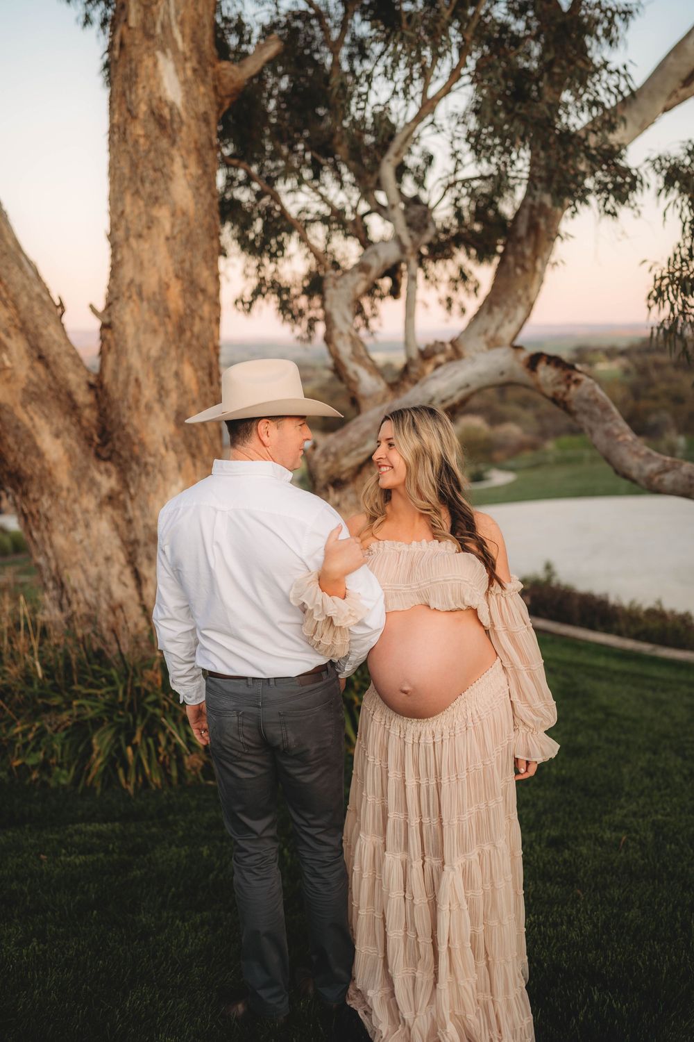 Pregnant woman in boho lace dress with partner in cowboy hat standing under eucalyptus tree at sunset.