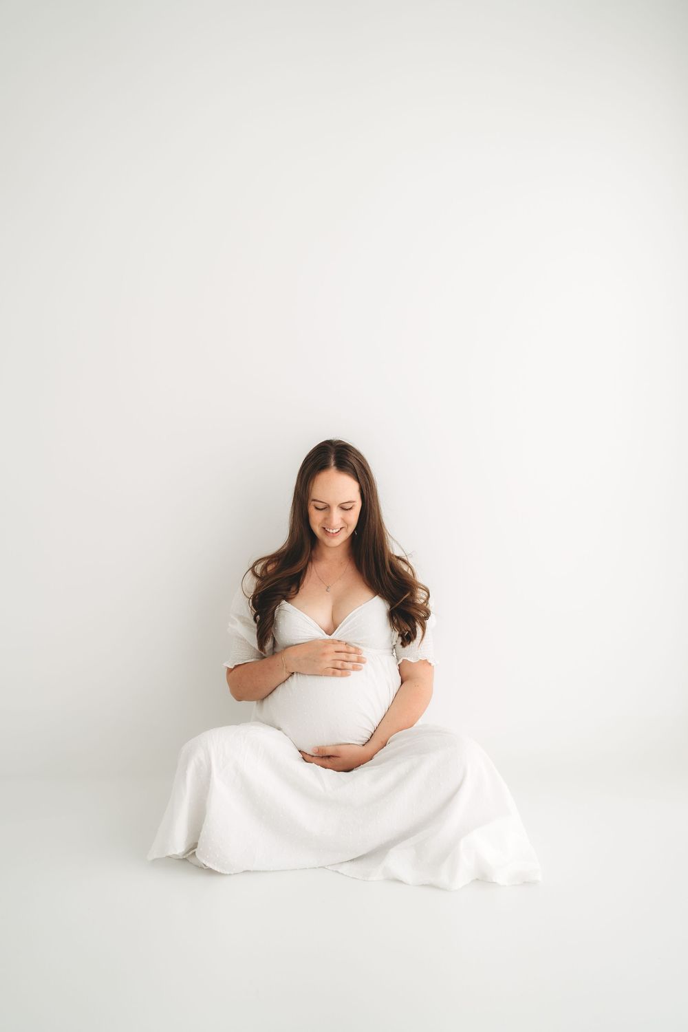 Pregnant woman in white dress sitting gracefully on floor, cradling baby bump in bright white studio.