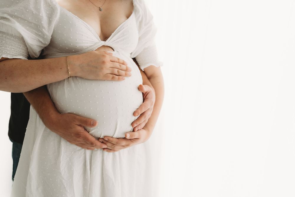 Close-up of couple tenderly holding pregnant belly together, wearing white clothing against light background.