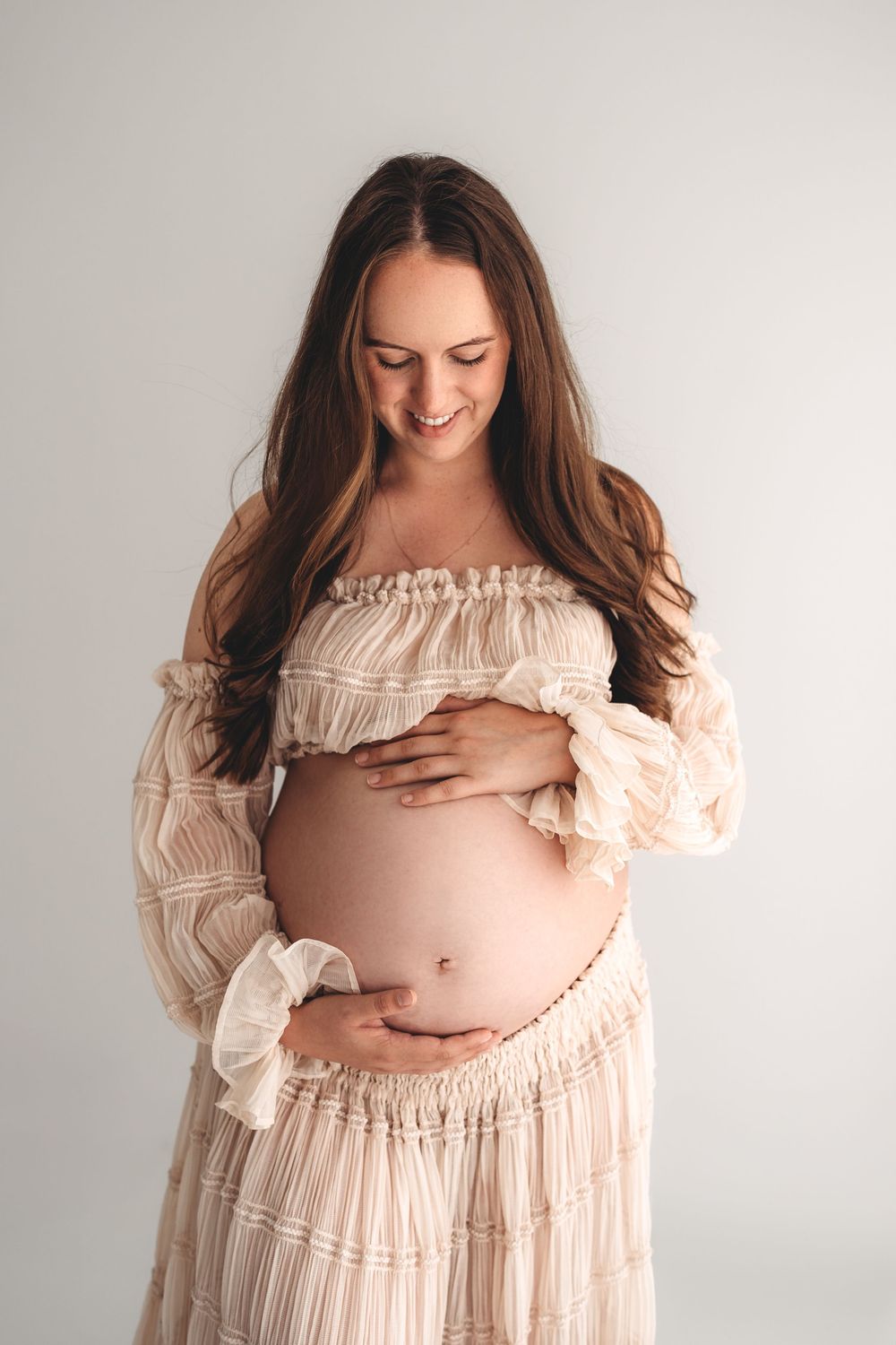 Smiling pregnant woman with long brown hair in boho off-shoulder dress gently holding bare baby bump.