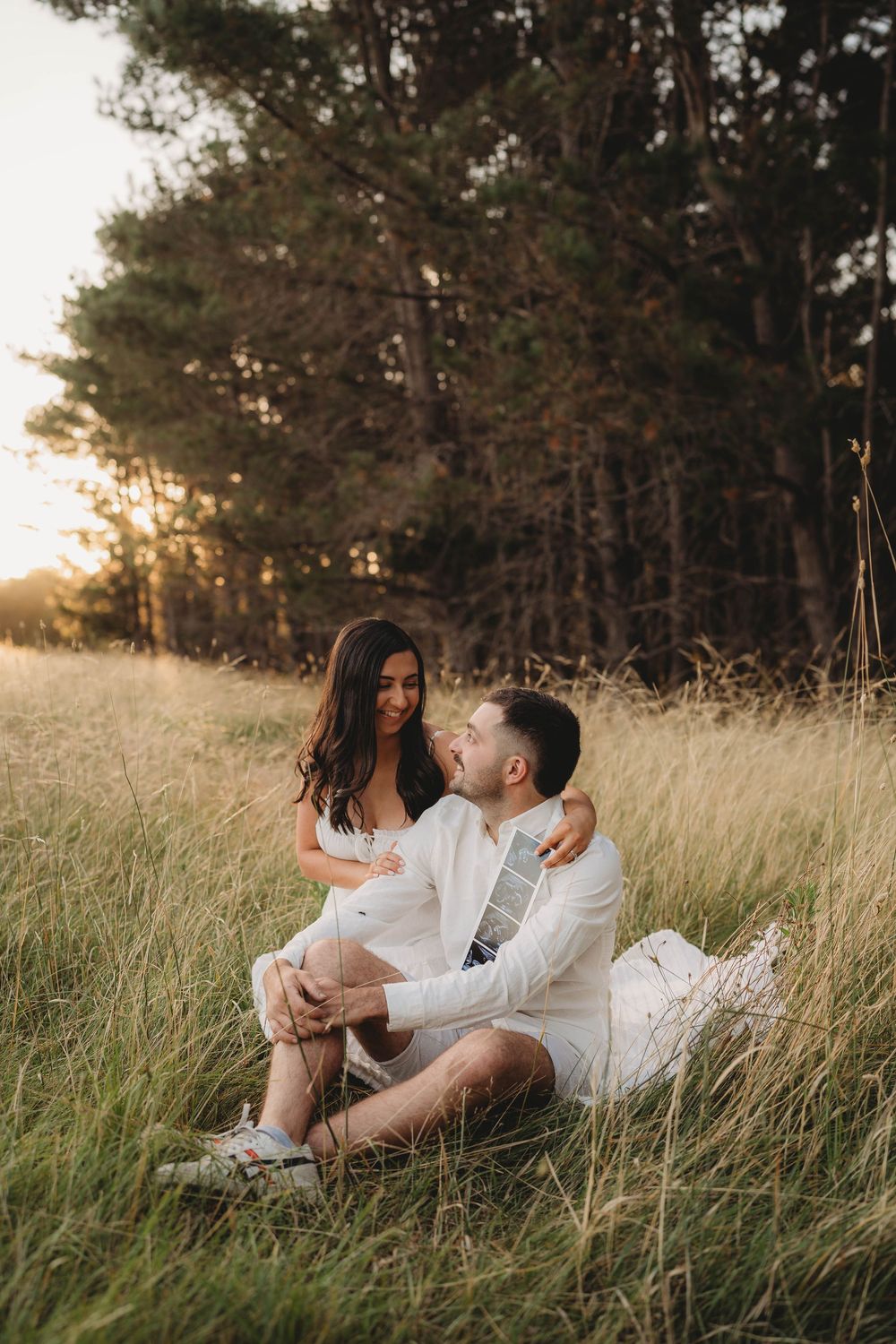 Couple sitting in golden grass field at sunset, man embracing pregnant woman in white dress near pine trees.