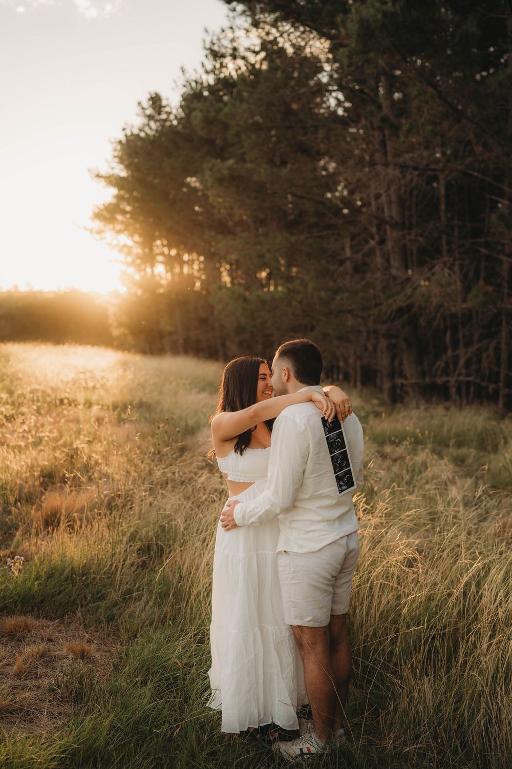Couple kissing at sunset in tall grass field near pine trees, woman holding ultrasound scan behind mans back.