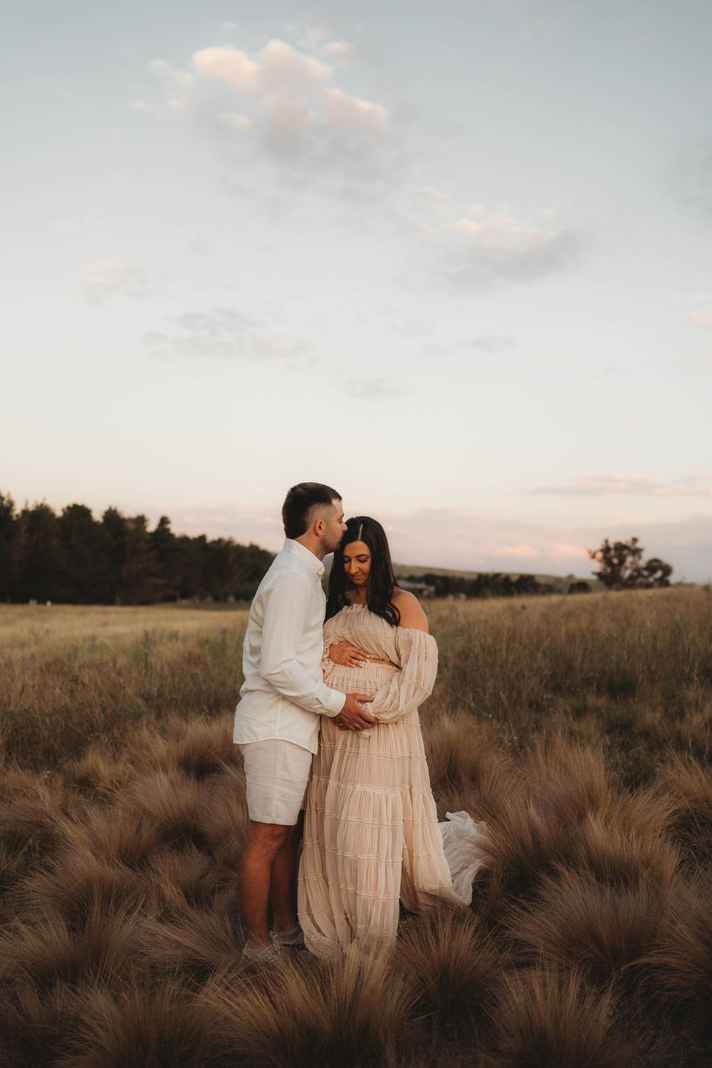 Couple standing tenderly in golden grass field at dusk, man embracing pregnant woman in blush ruffled gown.