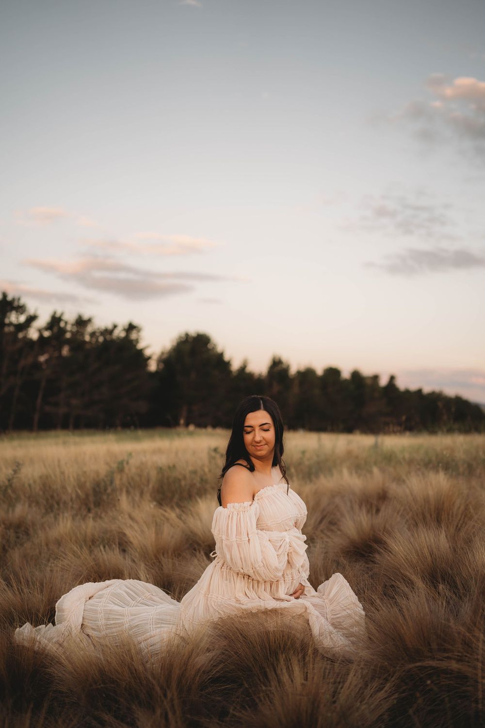 Pregnant woman in flowing off-shoulder blush gown sitting peacefully in golden tall grass field at sunset.