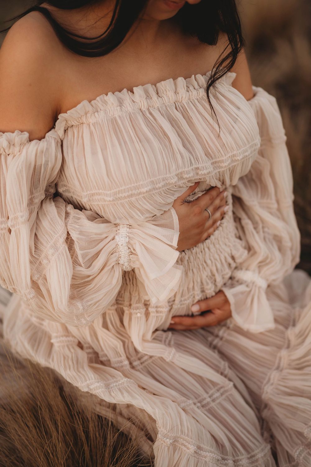 Close-up of pregnant woman in elegant off-shoulder ruffled blush gown cradling baby bump with ringed hands.