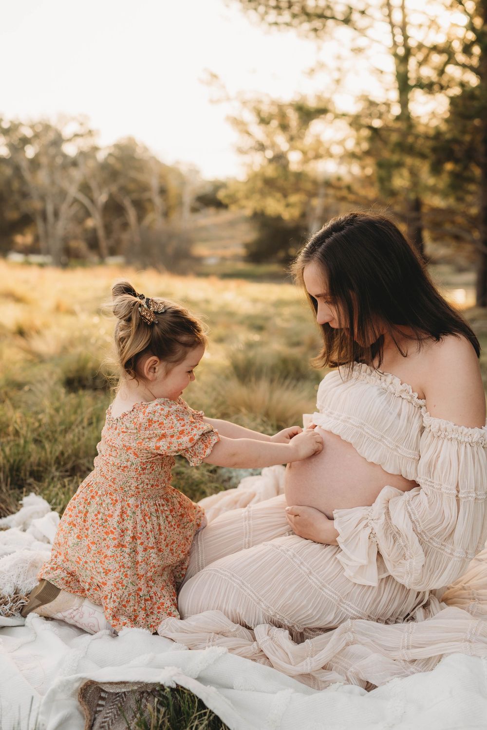 Pregnant woman in cream off-shoulder dress sitting outdoors with young girl in floral dress touching baby bump.