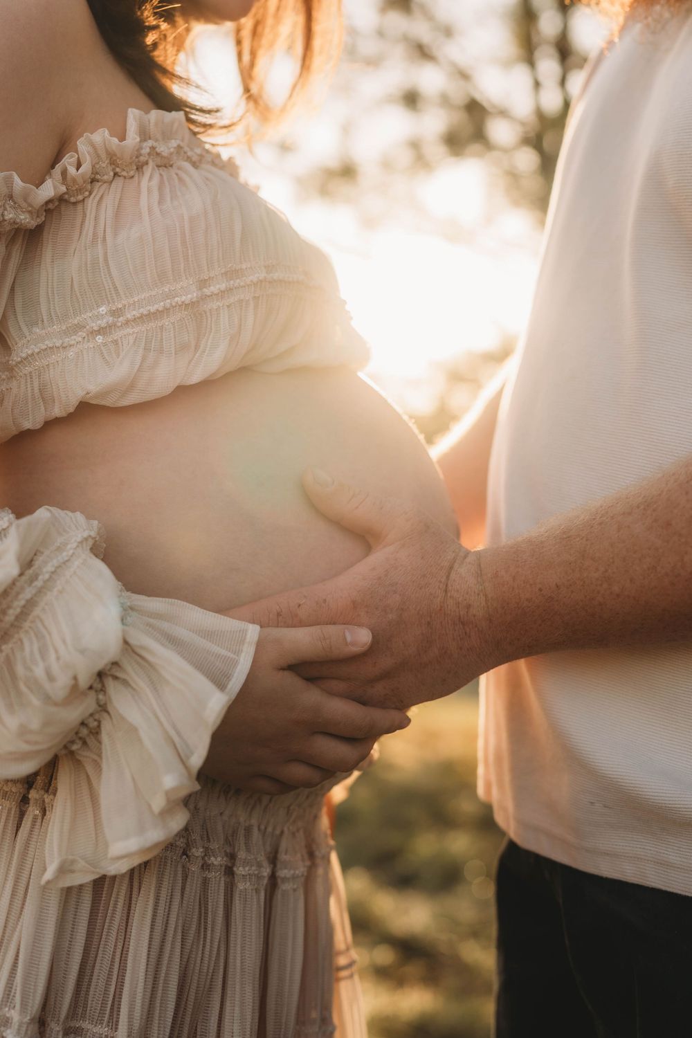 Close-up of couple holding pregnant belly together at golden sunset with warm lens flare in background.