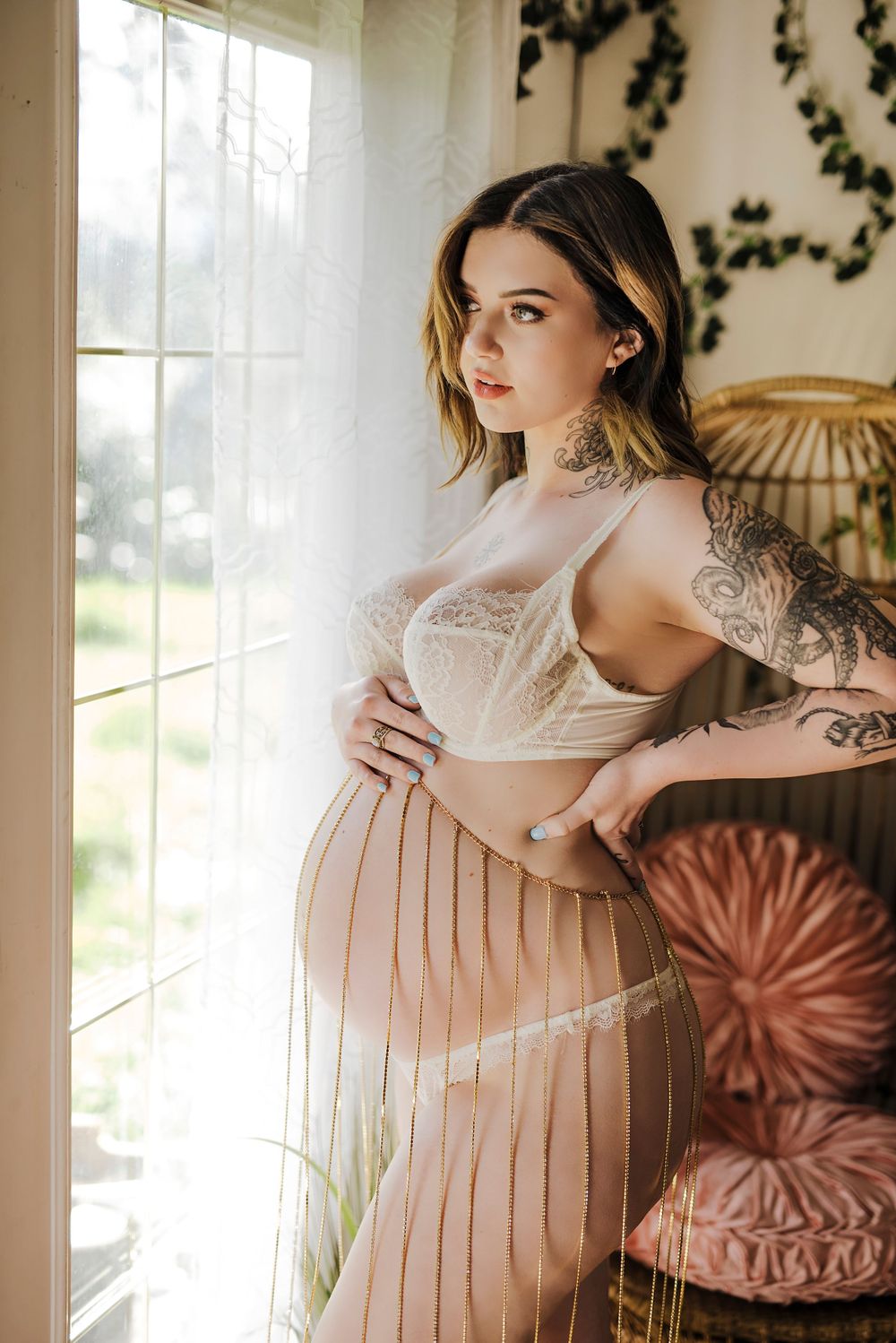 Pregnant woman in sheer lace lingerie, holding her belly while gazing thoughtfully out the window, bathed in soft natural light.
