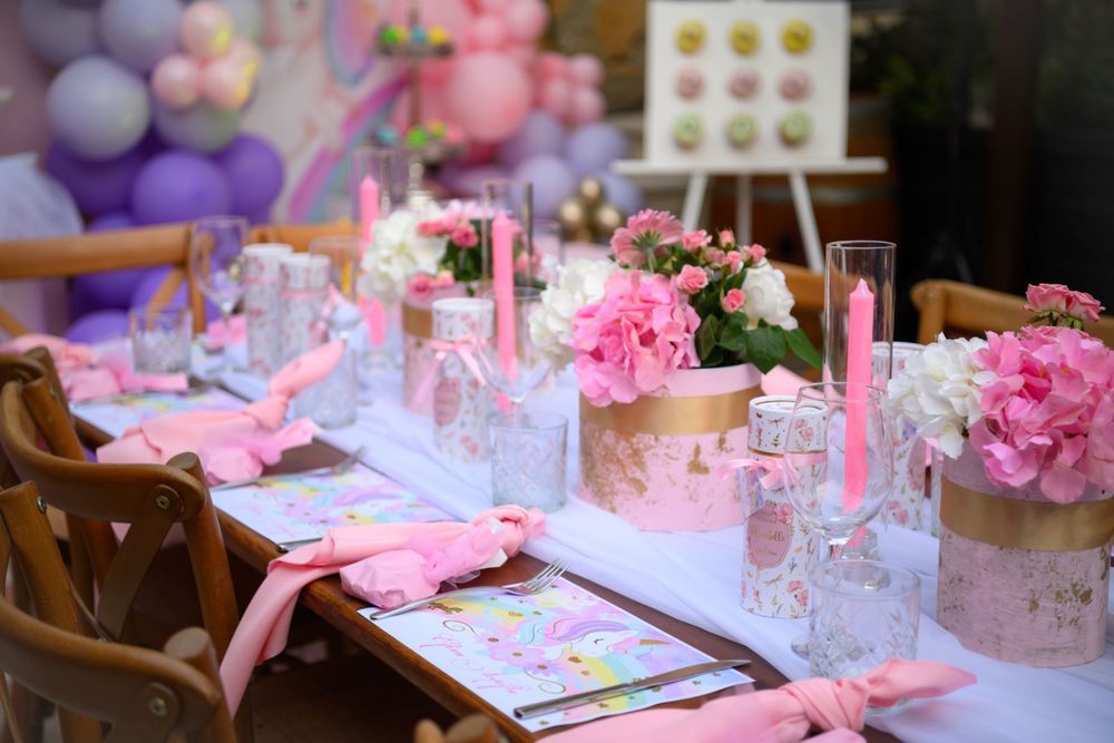 Pink-themed party table with floral arrangements and place settings under purple balloon decor.