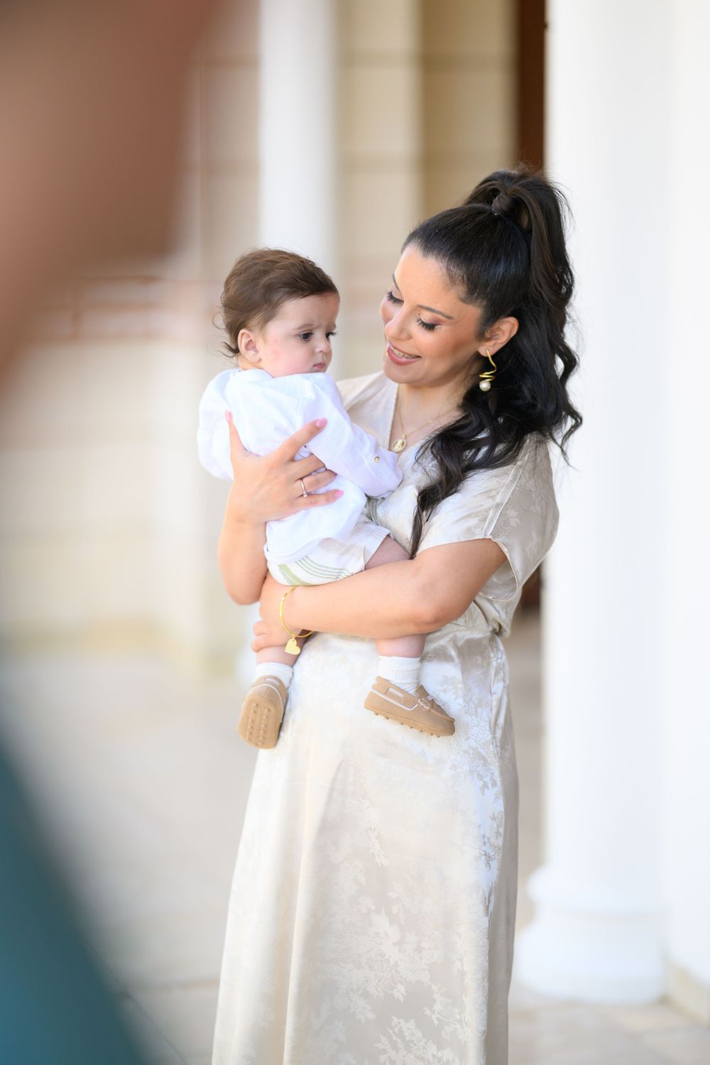 Parent in white dress holds baby in elegant architectural setting.