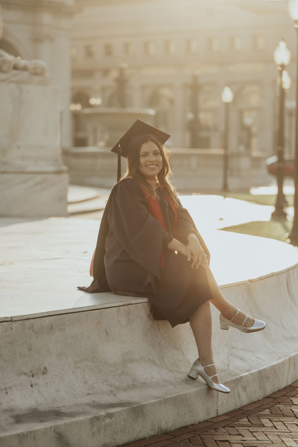 A graduate in cap and gown sits on concrete steps during golden hour, illuminated by warm sunlight and street lamps.
