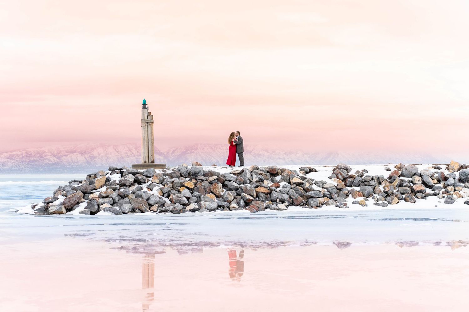 A dreamy pink sunset scene at a lighthouse with figures silhouetted on a rocky breakwater in Saratoga Springs, Utah.