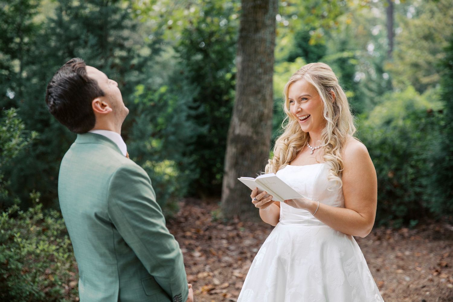 Bride in white dress reads vows to groom in sage green suit during first look at little river farms wedding in milton, georgia