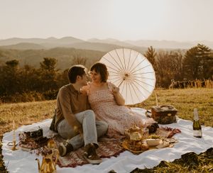 a couple enjoys a picnic on top of a hill using a parasol for shade from the sun as it sets over the Blueridge mountains of Asheville