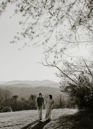 a monochrome shot of a couple walking up a hill with tree branches in the foreground and the Blueridge mountains of North Carolina behind