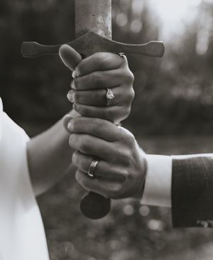 a bride and groom show off their rings in black and white as they hold a sword together