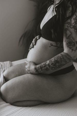 a pregnant woman kneeling on the bed holding her stomach with both hands and tattoos