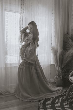 a monochrome shot of a mother to be in front of a window in a dress