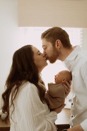 a mom and dad share a kiss while holding their infant child between them as light pours in from behind them