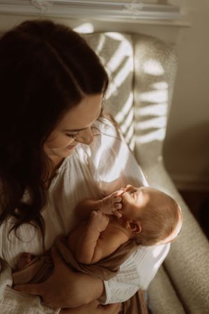 a mother sits in a beige chair as light from the windows and blinds makes a pattern while she looks down at her infant