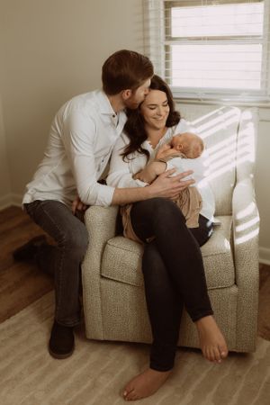 a man kneels beside a beige chair to kiss the mother of his child on the forehead as she holds her newborn baby and smiles down at him