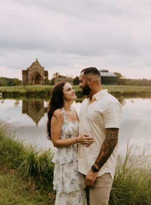 a couple smile at each other in front of a pond and wedding chapel