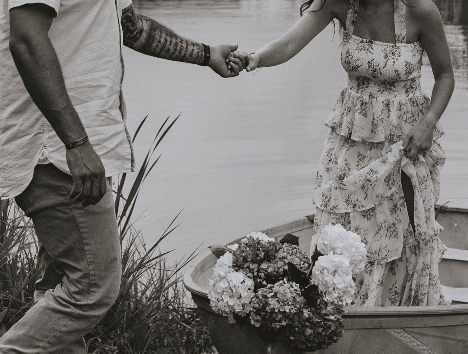 a man with a tattooed arm helps a woman in a floral dress out of a canoe that has a bouquet of flowers on it