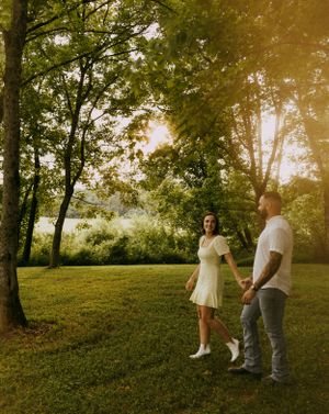 a man and woman walk hand in hand in a lush green yard filled with trees