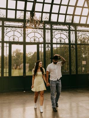 a man fixes his cowboy hat as he walks hand in hand with a woman in a yellow dress inside a glass house with chandeliers