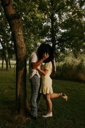 a man hides his kiss with a woman by using his cowboy hat they are leaned up against a tree and the woman is lifting her leg