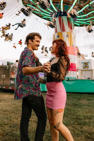 a couple in colorful clothing hold hands in front of the moving swings at the carnival in Western North Carolina near Asheville and Boone