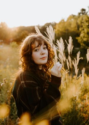 girl in sweater in golden light surrounded by tall grasses a girl in a sweater chews on a wheat grass surrounded by golden light and flowers