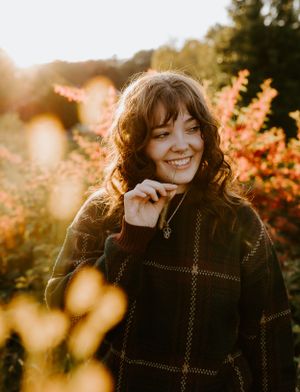 a girl in a sweater chews on a wheat grass surrounded by golden light and flowers in Western North Carolina near Asheville