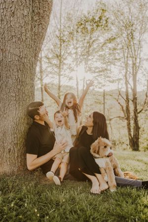 a young family with their dog sit at the base of a tree on tree and celebrate the joys of being together