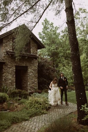 a bride and groom walk down a brick path in front of a stone house set in the Blueridge Mountains of Asheville