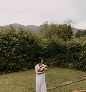 a mother in a white dress holds her infant son outside with the Blueridge Mountains of Waynesville in the background