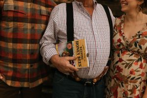 three people wearing patterned shirts the one in the middle is wearing suspenders and holding a book about beekeeping