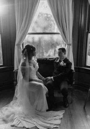 in monochrome a bride and groom sit together holding hands on a couch in front of the window that is letting light in
